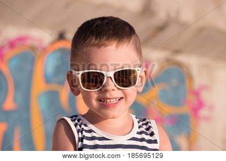 smiling small kid wearing sunglasses with sea sunset reflection in it and sailor shirt on graffiti background lit by warm summer light