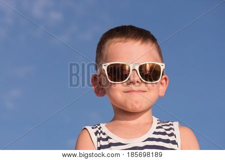 glad little boy wearing white sunglasses and sailor stripes shirt on blue sky background