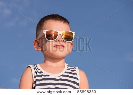 happy little boy wearing white sunglasses and sailor stripes shirt on blue sky background