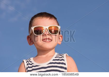 smiling little boy wearing white sunglasses with sea sunset reflection and sailor stripes singlet on blue sky background with copyspace