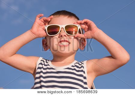 smiling little boy wearing white sunglasses with sea sunset reflection and sailor stripes vest showing teeth on blue sky background with copyspace