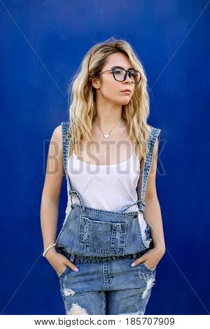 Young Stylish Blond Girl In Denim Overalls Made Of Denim And On A Blue Background