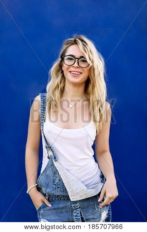 Young Stylish Blond Girl In Denim Overalls Made Of Denim And On A Blue Background
