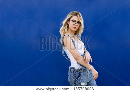 Young Stylish Blond Girl In Denim Overalls Made Of Denim And On A Blue Background