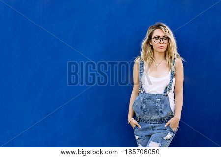 Young Stylish Blond Girl In Denim Overalls Made Of Denim And On A Blue Background