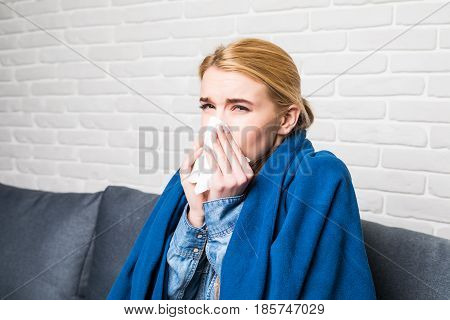 Blonde Woman Sneezing In A Tissue On Sofa In The Living Room