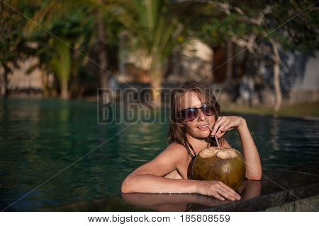 Horizontal portrait of sexy girl in sunglasses sitting in water with arms and coconut with straw on pool edge