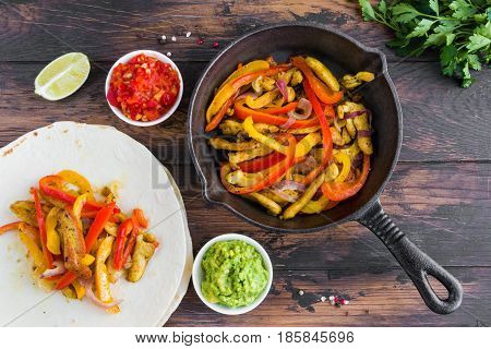 Chicken fajitas fillet roasted with bell peppers and red onion in a black skillet tortillas and traditional Mexican sauces salsa and guacamole. Wooden rustic table top view.