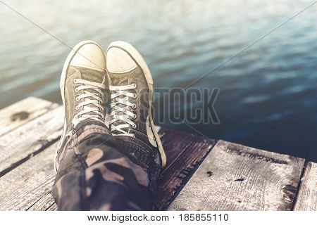Man with crossed legs relaxing on wooden riverbank pier casual young guy wearing sneakers sitting by the river in summer afternoon selective focus