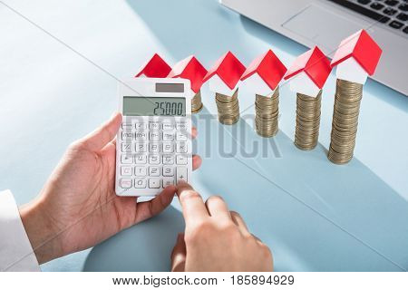 Person Showing Profit On Calculator With House Models On Coins Stack Over The Blue Desk