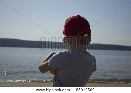A small child looks out at the water with the ball in hand