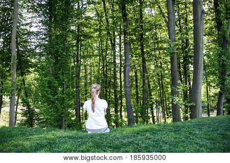 A girl in a white t-shirt sits and stares into the green forest. Rear view.