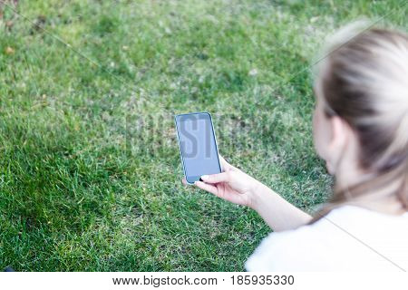 The phone in the girl's hand against the background of the green grass. The girl is writing a message in the park. Rear view.
