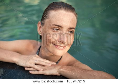 Horizontal close-up portrait of beautiful girl with sunspots and wet hair in water, arms on pool edge