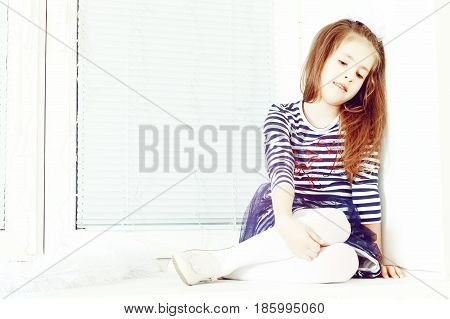 Little girl in blue striped dress and a white bow on her head.She sits at the window with sad eyes something lost in thought.Creative toning of a photograph.