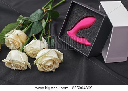 Only For Adults. Close Up Photo Of Pink Sex Toy In A Gift Box And White Roses Against Of Black Silk