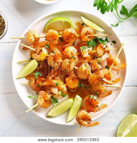 Close Up Of Prawns Skewers With Greens, Species, Lime And Sauce On White Plate Over Wooden Table. Gr