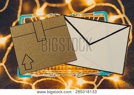 Shopping Cart With Cardboard Parcel And Email Envelope On Concrete Desk Surrounded By Fairy Lights, 