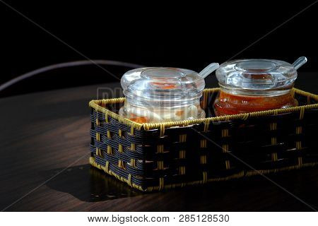 Bottles Of Seasoning With Spoons In Rattan Box On Wooden Dining Table On Dark Background
