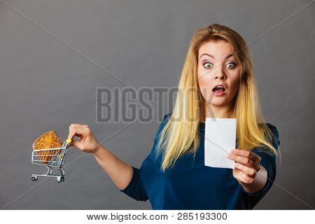 Shocked Woman Holding Shopping Basket With Bread Looking At Bill Receipt Being Scared Of Huge Prices