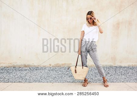 Hipster girl wearing blank white t-shirt and grey pants holding wicker handbag posing against street wall, blank mockup for tshirt print store