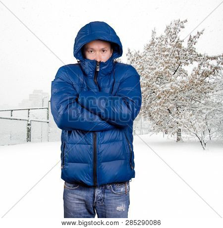 Sad Asian man in blue down padded coat, against snowy winter background. Man outdoors in warm winter clothes. Snow and cold