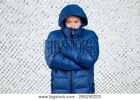 Sad Asian man in blue down padded coat, against snowy winter background. Man outdoors in warm winter clothes. Snow and cold