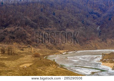 Winter Landscape Of Frozen River In Mountain Valley With Small Concrete Building On Riverbank.