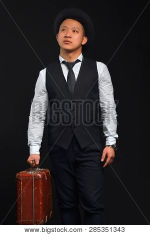 Asian Man In A Hat And A Suit With A Brown Suitcase Looks Up, Thoughtful, Photo In Full Swing