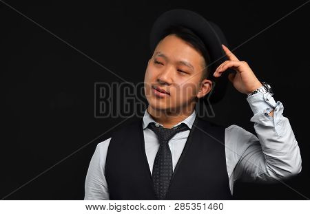 An Asian Man Wearing A Black Hat, Shirt And Tie, Stands On A Black Background, Looks Aside And Touch