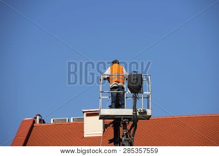 The Worker In Overalls Works At Height In A Building Mechanical Lifting Basket. Repair And Construct