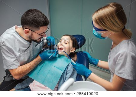 Male Dentist Check Up Girls Teeth With Dentists Tools. Female Helper Stand Beside. Girl Sit In Denta