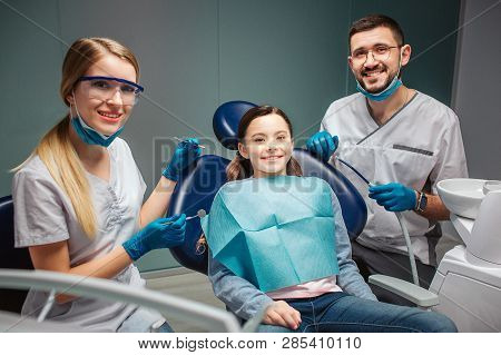 Cheerful Nice Young Man Woman Detists Sit And Pose With Child In Dental Chair. Adults Hold Tools. Si