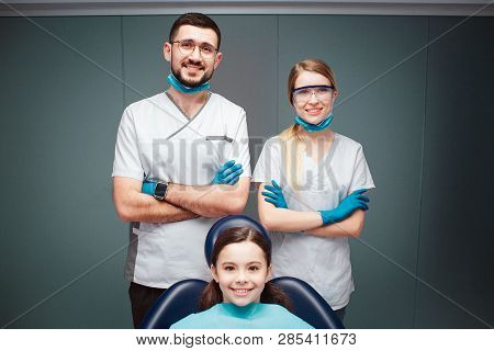 Nice Positive Male And Female Dentist With Girl In Dental Chair. They Look Straight And Smile. Adult