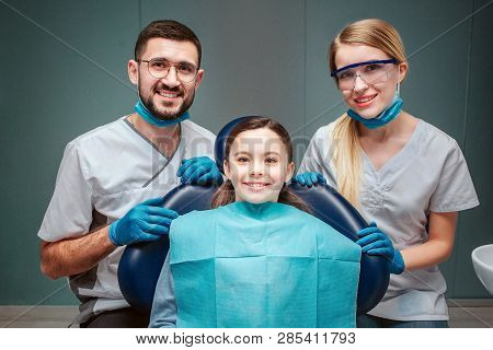 Positive Cheerful Male And Female Dentist With Girl Patient. They Look Straight Together And Smile. 