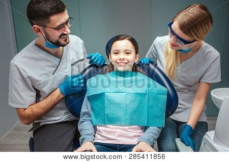 Careful Positive Male And Female Dentist Look At Kid Patient And Smile. Girl Sit In Dental Chair. Sh