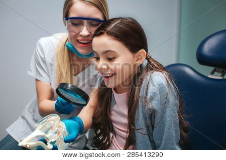 Amazed Girl Sit In Dental Chair In Room. She Look At Artificial Jaw With Teeth. Female Dentis Hold L