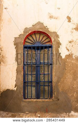 Wall Of An Old House With Peeled Off Paining Of The Facade. Red Window Arch, Blue Grating. Street Of