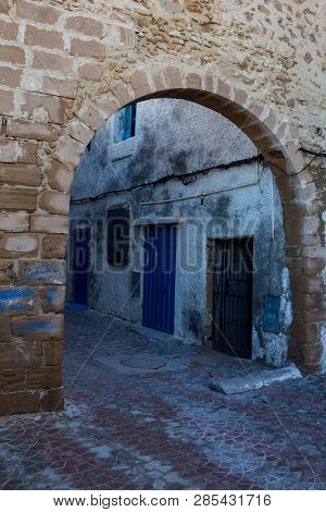 Dark Morning Narrow Street With An Arch City Gate, Made Of Stone Bricks. Colorful Paving. Blue Doors