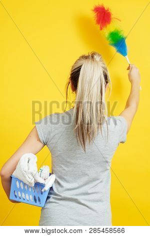 Dust Cleaning. Woman With Feather Duster And Basket Of Cloths On Yellow Background. Backview.