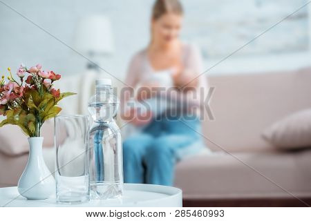 Close-up View Of Glass, Bottle Of Water, Flowers In Vase And Mother Breastfeeding Baby Behind At Hom