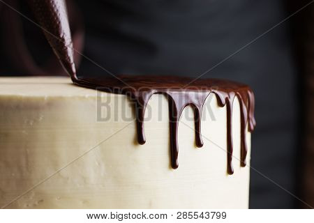 A Confectioner Squeezes Liquid Chocolate From A Pastry Bag On To A White Cream Biscuit Cake. The Con