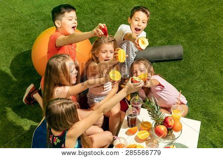 Kids Fashion Concept. The Group Of Teen Boys And Girls Sitting On Green Grass At Park. Children Colo