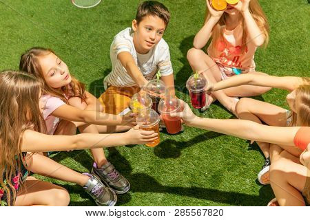 Kids Fashion Concept. The Group Of Teen Boys And Girls Sitting On Green Grass At Park. Children Colo