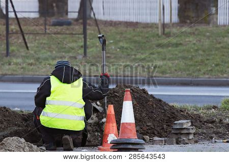 Worker Repairs The Road Bending Over Dug Hole. A Man With A Shovel Inspects The Moat. Overalls With 