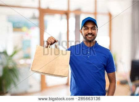 takeaway service and people concept - happy indian delivery man food in paper bag in blue uniform over office background