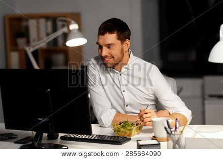 business, overwork, deadline and people concept - smiling businessman at computer eating vegetable salad at night office