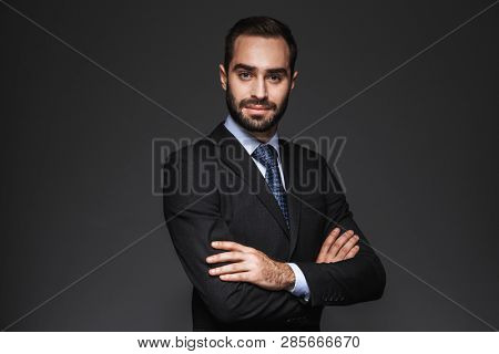 Portrait of a confident handsome businessman wearing suit isolated over black background, arms folded