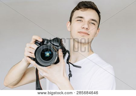 A Young Man Holds A Photo Camera In His Hand And Looks Straight. Isolated Gray Background
