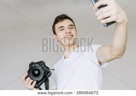 A Young Man Holds A Photo Camera In His Hand And Makes A Selfie By Phone. Isolated Gray Background